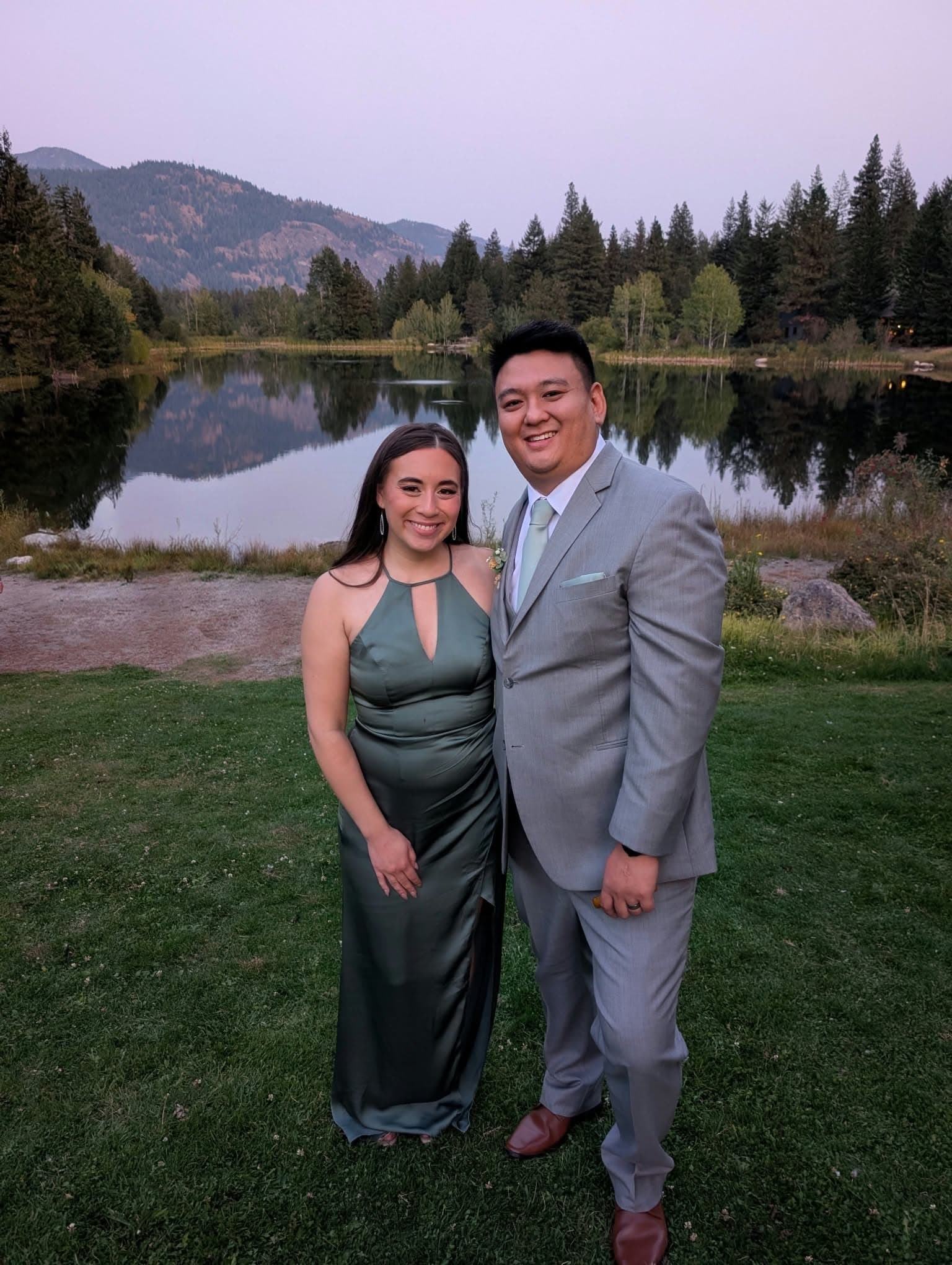 Nam Nguyen in a grey suit, smiling beside a woman in a green dress, in front of a mountain lake at dusk.
